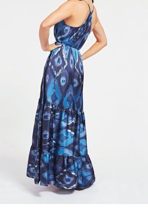 Kvinde iført en blå mønstret maxikjole uden ærmer med lagdelte flæser, poserer med hænderne på hofterne mod en ensfarvet hvid baggrund.