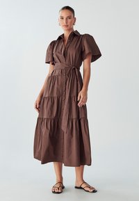 Braunes, gestuftes Kleid aus Baumwolle mit kurzen Puffärmeln, Knopfverschluss und Taillengürtel. Kombiniert mit schwarzen flachen Sandalen.