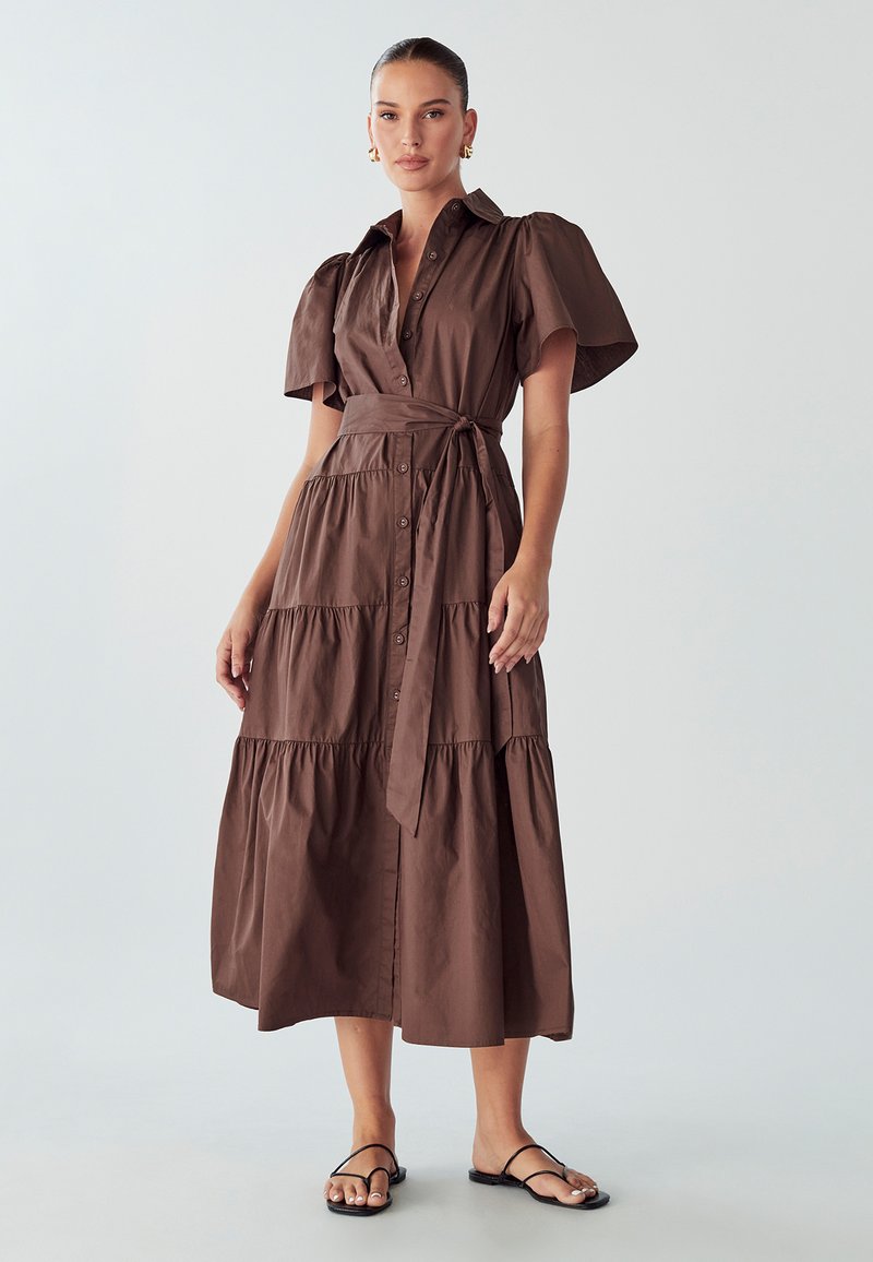 Braunes, gestuftes Kleid aus Baumwolle mit kurzen Puffärmeln, Knopfverschluss und Taillengürtel. Kombiniert mit schwarzen flachen Sandalen.