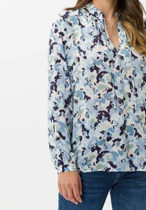 Kobieta ubrana w bluzkę z długim rękawem w abstrakcyjny niebiesko-czarno-biały kwiatowy wzór, w połączeniu z niebieskimi dżinsami, stojąca z jedną ręką luźno opuszczoną.