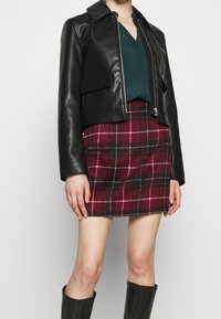 Chaqueta corta de cuero negra con un cuello abotonado, combinada con una falda de cuadros roja y negra. La modelo lleva botas negras hasta la rodilla.