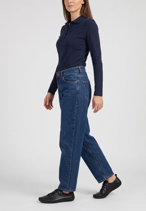 Kvinde iført en navy langærmet polo, blå straight-leg jeans og sorte snøresko til hverdagsbrug stående på en ensfarvet lys baggrund.
