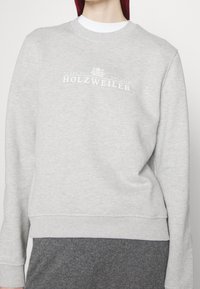 Šedá bavlněná mikina s kulatým výstřihem, opatřená žebrovanými manžetami a spodním lemem. Na hrudi se nachází bíle tištěné logo "HOLZWEILER". Měkká textura.