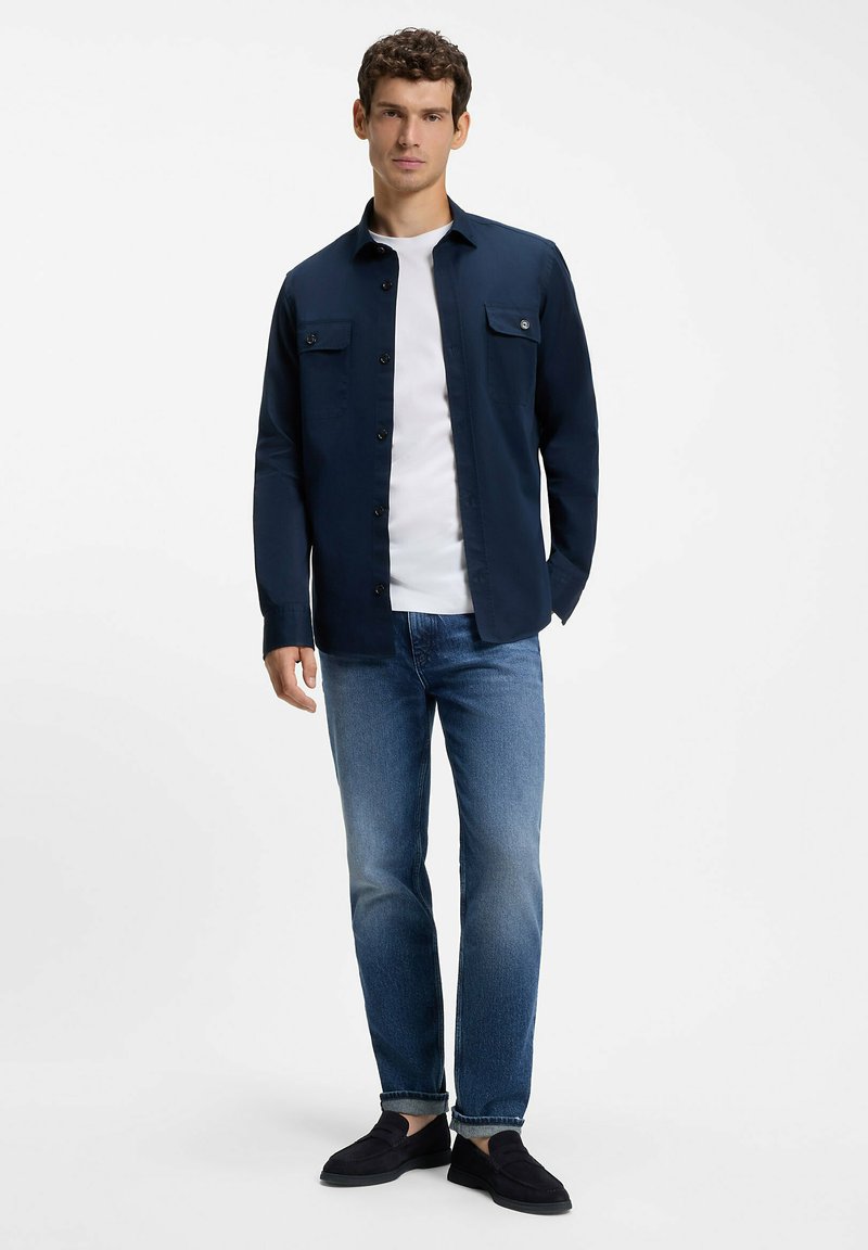 Homme debout portant une chemise bleu marine à boutons par-dessus un t-shirt blanc, un jean bleu et des chaussures noires sans lacets sur un fond blanc uni.