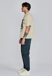Beige graphic t-shirt met een zwarte print, gecombineerd met donkergroene cargobroeken. Het model draagt witte sneakers met textuurveters.