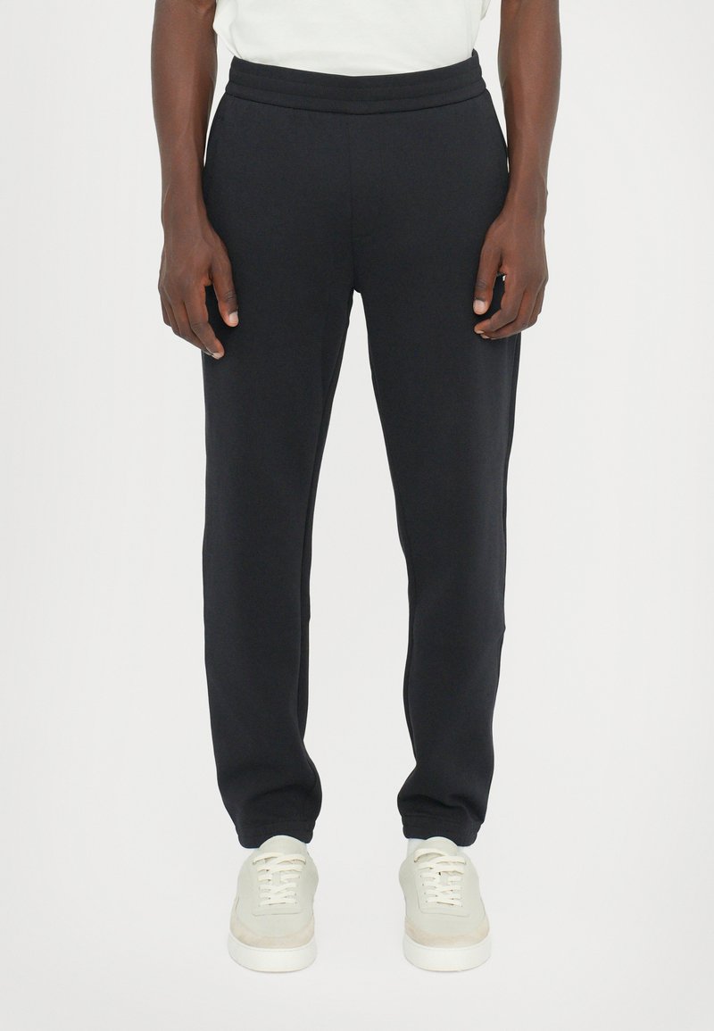 Black sweatpants with a drawstring waist, tapered leg, soft texture, and side pockets, paired with light-colored sneakers.