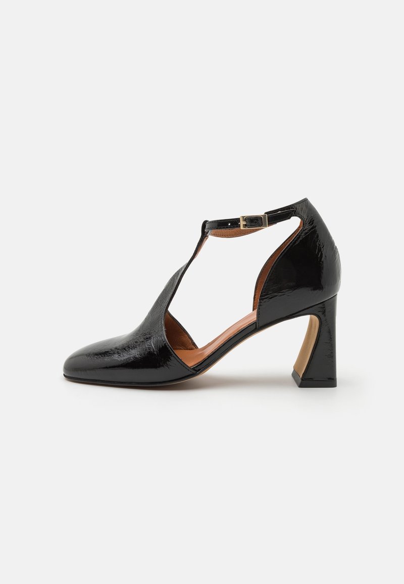 Chaussure en cuir verni noir à talon moyen, avec bride en T et fermeture à boucle, dotée d'un design latéral ouvert et d'un bout arrondi.
