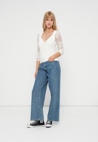 Biely čipkovaný top s dlhým rukávom a V-výstrihom, kombinovaný s širokými modrými džínsami a čiernymi teniskami s bielymi akcentmi.