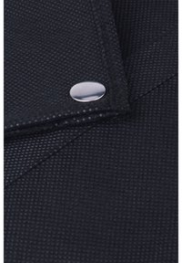 Czarna fakturowana tkanina z wyraźnym srebrnym zatrzaskiem, z charakterystycznym trójkątnym klapem i widocznymi detalami szwów.