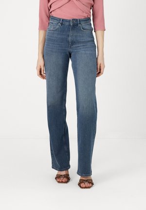 Femme de la taille aux pieds portant un jean bleu taille haute à jambes droites, un haut rose à manches trois-quarts et des sandales marron ornées de clous.