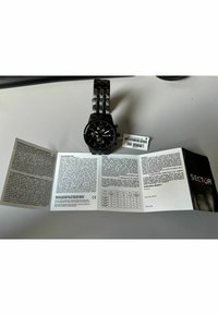 Orologio in acciaio inossidabile nero con quadrante rotondo, tre sotto-quadranti, bracciale a maglie e manuale utente spiegato accanto, con testo in bianco e nero.