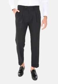 Pantaloni neri a pieghe con vestibilità su misura, chiusura a due bottoni, tasche laterali e orli risvoltati, abbinati a mocassini neri.