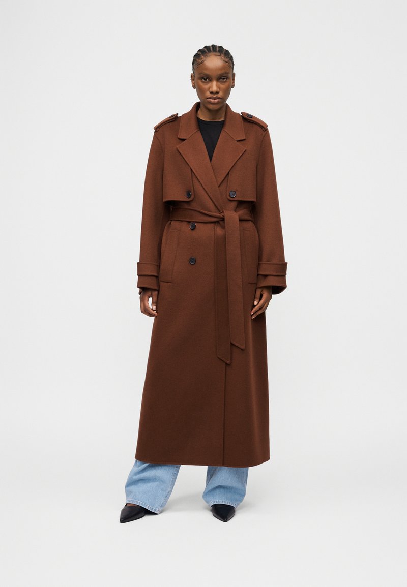 Γυναίκα που φοράει μακρύ καφέ διπλόμπουτο παλτό με ζώνη πάνω από ανοιχτό μπλε τζιν και μαύρα μυτερά παπούτσια, στέκεται μπροστά σε απλό φόντο.