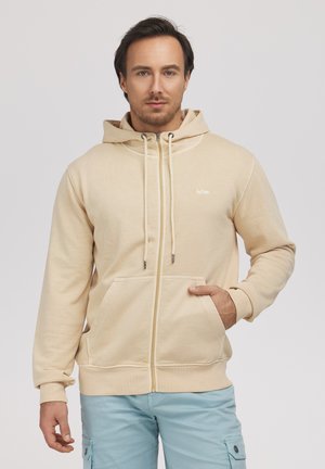 Mężczyzna w beżowej bluzie na zamek z kapturem i ściągaczami oraz jasnoniebieskich spodniach cargo, pozuje z jedną ręką w kieszeni na białym tle.