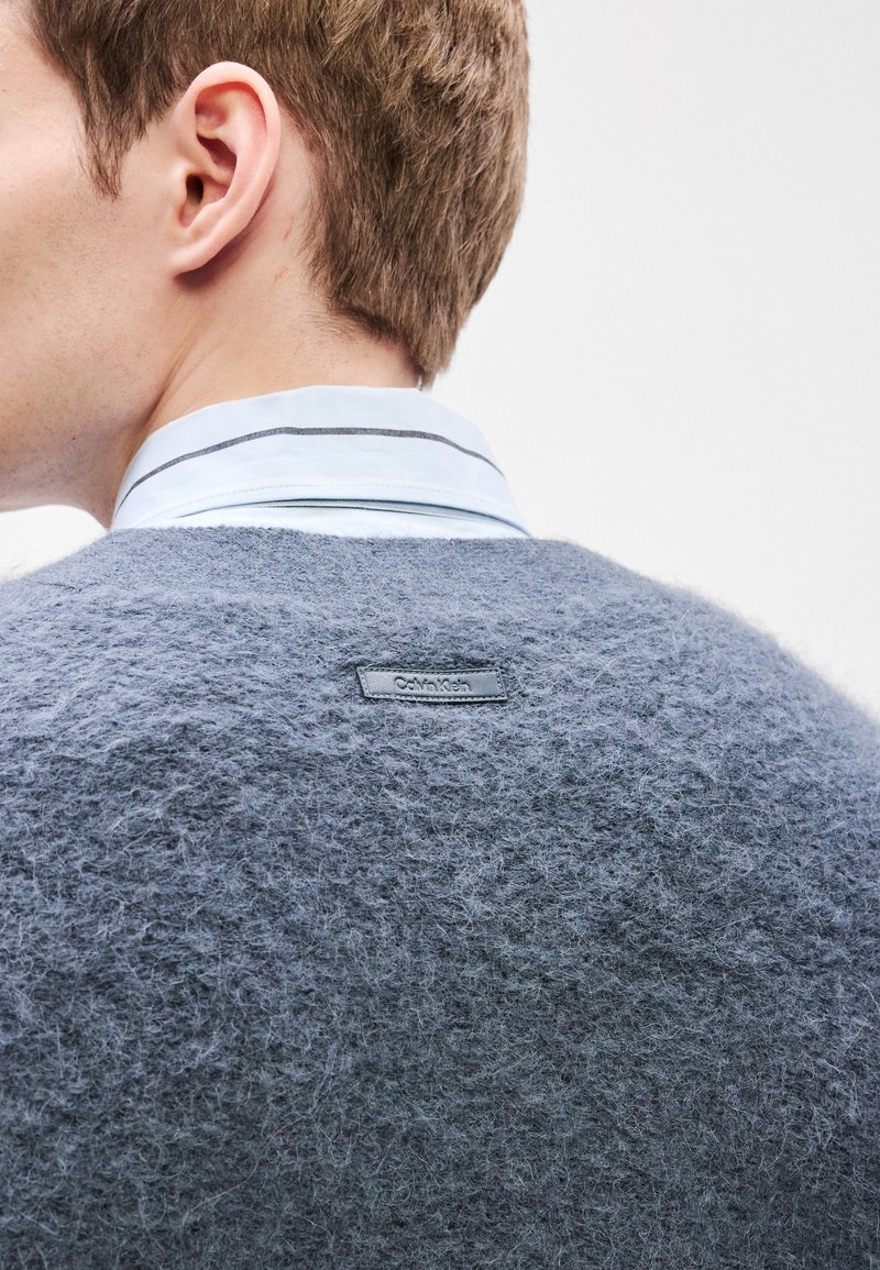 Grey knitted sweater with a fuzzy texture, featuring a black Calvin Klein logo tab at the back and a light blue collared shirt underneath.