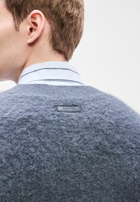 Grey knitted sweater with a fuzzy texture, featuring a black Calvin Klein logo tab at the back and a light blue collared shirt underneath.