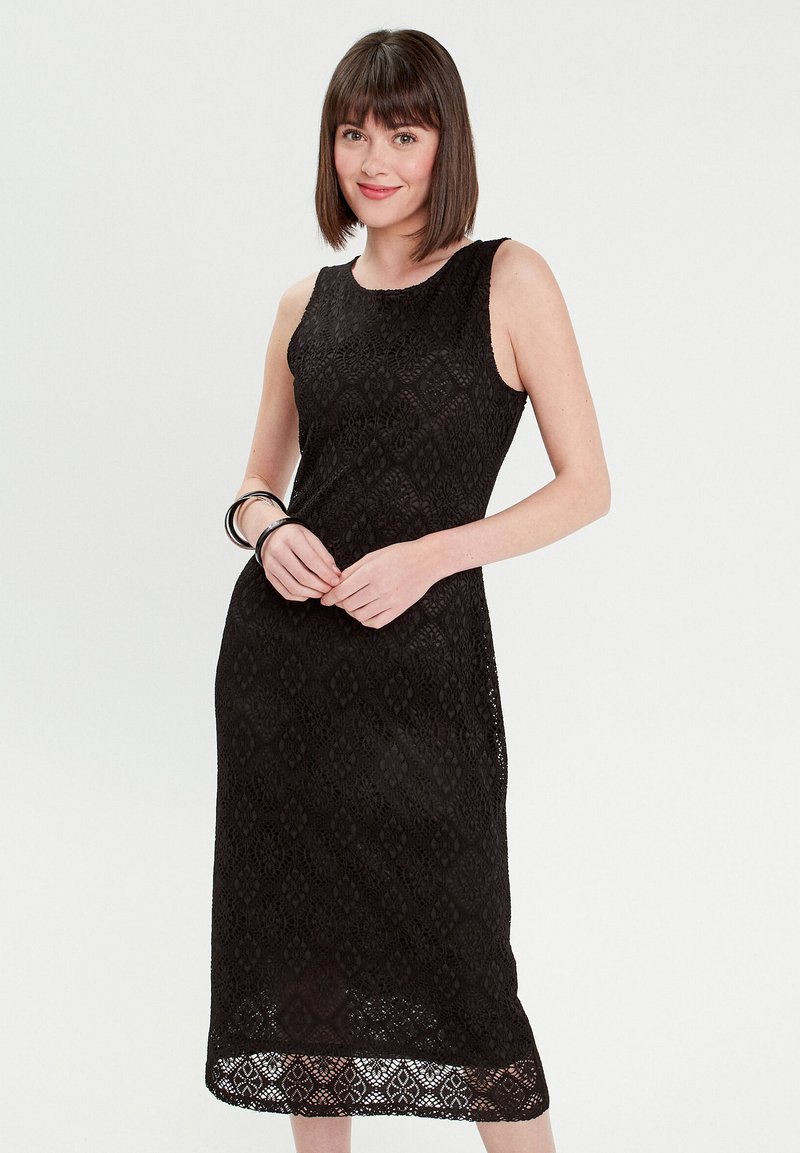 Robe en dentelle noire avec un design sans manches, motif en losanges, longueur au genou et finition texturée. Portée avec des bracelets en argent au poignet.