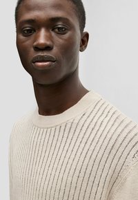 Beige knit sweater with horizontal ribbing pattern, round neckline, and a soft texture; features thin, evenly spaced stripes throughout.