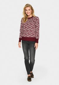 Pull maroon avec un motif abstrait blanc, col et poignets côtelés. Associé à un jean sombre et des chaussures en cuir frangé marron.