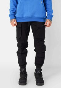 Zwarte cargobroek met meerdere zakken, ribgebreide boorden en een trekkoord in de taille, gecombineerd met zwarte sneakers met mesh- en suède-accenten.