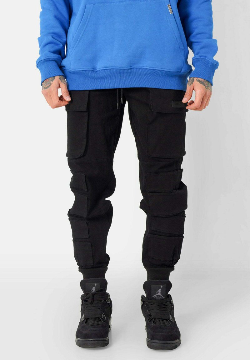 Zwarte cargobroek met meerdere zakken, ribgebreide boorden en een trekkoord in de taille, gecombineerd met zwarte sneakers met mesh- en suède-accenten.