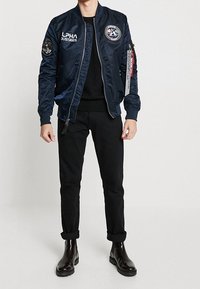 Chaqueta bomber azul marino con acabado de satén suave, con parches y detalle de cremallera. Pantalones negros ajustados y botines brillantes hasta el tobillo.