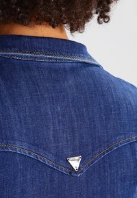 Denimjacka med en spetsig yoke-design, triangel-logotyp på baksidan och kontrasterande gul sömnad på mörkblått tyg.