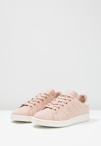 Ett par ljusrosa mocka lågskor med vita sulor och snören, perforerade sidosträck, på en vit yta med grå bakgrund.