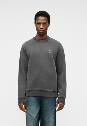 Młody mężczyzna w ciemnoszarej bluzie z małym białym napisem na piersi oraz w niebieskich dżinsach na jednolitym jasnym tle.