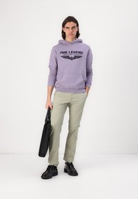 Svetlofialová mikina s kapucňou a čiernym grafickým potlačou, v kombinácii so svetlozelenými nohavicami a čiernymi topánkami. Drží čiernu tašku.