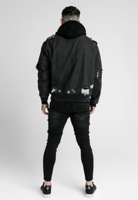 Czarna postarzana kurtka jeansowa z postrzępionymi krawędziami, noszona na czarnej bluzie z kapturem. Stylizację dopełniają obcisłe czarne jeansy i czarne buty.