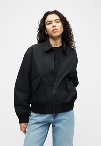 Νεαρή γυναίκα με σγουρά μαλλιά, φορώντας μαύρο μπουφάν bomber και ανοιχτό μπλε τζιν, μπροστά σε απλό λευκό φόντο.