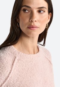 Pull en maille rose clair, texturé, avec un col rond et des manches raglan, confectionné dans un matériau doux et duveteux. Design simple sans motifs.