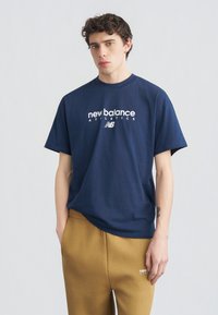 Νεαρός άνδρας που φοράει μπλε μπλούζα New Balance και μουσταρδί φόρμα, στέκεται μπροστά σε απλό φωτεινό φόντο.