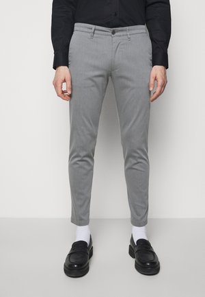Mand iført slim fit grå bukser, hvide sokker, sorte loafers og en sort skjorte med knapper, der står mod en ensfarvet lys baggrund.