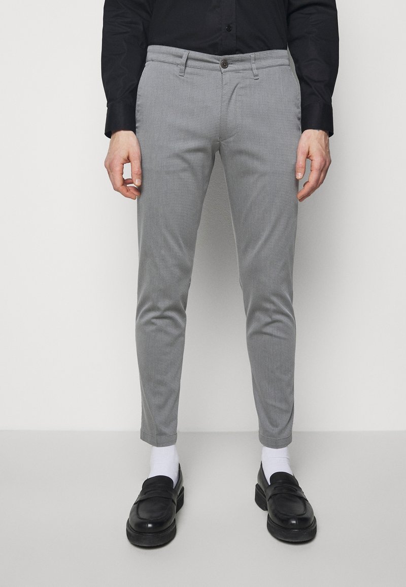 Homme portant un pantalon gris slim, des chaussettes blanches, des mocassins noirs et une chemise noire boutonnée, debout devant un fond clair uni.
