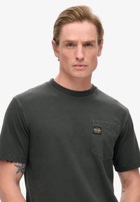 T-shirt a maniche corte verde scuro con scollatura rotonda, dotato di una tasca sul petto e dettagli di cucitura a contrasto. Tessuto morbido.
