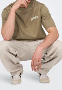 Oliwkowo-zielona koszulka z krótkim rękawem z nadrukiem "MONT" w kontrastowej czcionce, zestawiona z jasnobeżowymi spodniami i białymi trampkami.