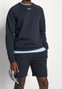 Uomo che indossa una felpa nera Under Armour e pantaloni corti, con una corda per saltare nera nella mano destra.