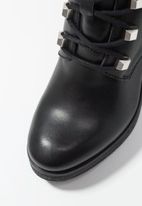 Botín negro de cuero hasta el tobillo con punta redonda, presenta un diseño de cordones y acentos metálicos cuadrados cerca de la parte superior. Textura suave.