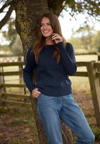 Suéter de punto trenzado en color marino con puños y dobladillo acanalados, combinado con unos vaqueros de corte relajado en azul claro. El modelo está de pie junto a un árbol.