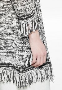 Czarny i biały dzianinowy ponczo z frędzlami, z teksturowanym wzorem i kontrastującymi paskami przy mankietach.