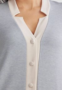 Ljust blå plagg med en beige krage, med ett V-halsdesign och fyra matchande tygklädda knappar framtill. Slät textur.