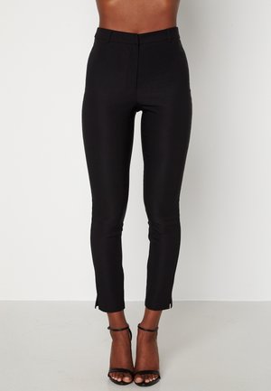 Mujer con pantalones negros ajustados de tiro alto y sandalias negras de tacón, de pie frente a un fondo blanco liso.