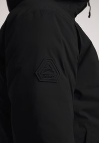 Giacca nera con una texture liscia e una toppa in gomma esagonale sulla manica con il nome "JACKIT". Design minimalista.