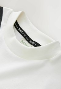 Gros plan sur le col ras du cou d'un T-shirt blanc avec une étiquette de marque noire indiquant "Baker by Ted Baker."