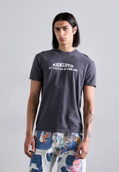 Gray cotton t-shirt featuring "KIDSUPER IT'S ALL UP IN THE AIR" text in white. Paired with textured, multicolored patchwork denim jeans.