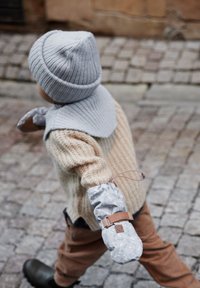 Kind, das eine hellgraue Mütze, einen beigen Strickpullover und braune Cordhosen trägt, hält einen grauen, gemusterten Handschuh mit Lederband.