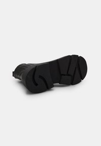 Suela de bota de goma negra con agarre texturizado, que presenta un diseño grueso y un detalle estampado a lo largo del borde. Tacón plano y puntera redondeada.