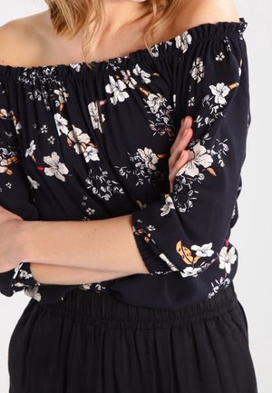 Frau trägt schwarze schulterfreie Bluse mit floralem Muster, elastischem Ausschnitt und Ärmeln, kombiniert mit einem schwarzen, hoch taillierten Rock oder Hose.